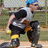 Animated icon of a character with blue hair, white shirt, black pants, and yellow knee pads, performing a side-to-side motion against a blurred outdoor background.