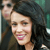 A static pixelated image shows a person with dark curly hair and a light skin tone smiling, wearing a dark top against a blurred background with green and brown tones.