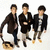 Three young men in dark jackets and light pants stand side-by-side on a white background, with one holding a guitar case.