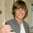 A pixelated image of a person with brown hair giving a thumbs-up gesture, wearing a gray jacket over a white shirt against a light background.