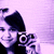 A person with dark hair holds a small camera, smiling against a light purple background.