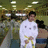 A person in a white karate uniform with a yellow belt stands in a gymnasium with blurred figures and overhead lights in the background.