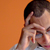 Pixelated close-up of a person with dark hair against an orange background, showing partial facial features and a light-colored garment.