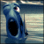 Blue cartoon character with large eyes and a black oval mouth, standing against a striped background.