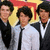 A group of six young men in formal attire pose against a yellow and red background, with three additional figures visible below.