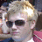 Static pixelated image of a person with light-colored hair, dark sunglasses, and a pink garment visible on the right side.