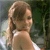 A pixelated image of a person with light brown hair tied back, wearing a white sleeveless top, standing against a blurred green and gray background.