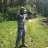 Pixelated static image of a figure standing in an outdoor setting with green grass and blurred foliage background.