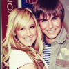 Two people with light brown hair pose closely together, one in a white top and the other in a striped shirt, against a red and blue background with partial text.