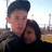 Two people embrace outdoors; one wears a dark cap and jacket, the other has dark hair and a dark top, with a bright sky and distant structures in the background.