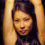 Pixelated portrait of a person with long dark hair against a warm-toned background, showing closed eyes and a neutral expression.