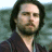Pixelated portrait of a person with dark hair and beard wearing dark clothing against a blurred greenish background.