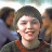 Pixelated static image of a person with dark hair and light skin wearing an orange-accented top, surrounded by blurred background figures in muted tones.