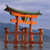 A pixelated orange torii gate with a green-tiled roof stands on a wooden platform over water under a light blue sky.