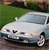 A silver sedan with visible headlights and wheels is positioned on a paved surface, with green grass and blurred foliage in the background.