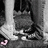 A grayscale pixelated image shows a figure in dark clothing with light-colored shoes, standing near vertical structures and a small pink shape on the ground.