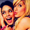 Two people pose closely together; one has dark hair and wears a pink beaded bracelet, the other has blonde hair and a light-colored top.
