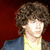 Pixelated image of a person with curly brown hair wearing a blue jacket over a yellow shirt, holding an acoustic guitar against a red background.