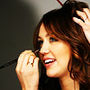 A close-up of a person with brown hair holding a microphone near their mouth, hand raised to hair, wearing a dark top against a gray background.
