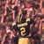 Two identical figures in dark uniforms with yellow accents stand side-by-side against a blurred crowd background, each holding a golden trophy-like object.