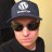 A pixelated close-up of a person wearing a dark cap with a white logo and dark sunglasses against a light background.