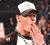 Two figures in white caps and dark clothing, one covering face with hand, the other holding a microphone against blurred background.