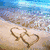 A split-screen animation showing beach sand with heart-shaped writing, ocean waves, and a dolphin leaping at sunset.