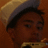 A pixelated close-up of a person wearing a light-colored hat with dark brim, holding an object near their mouth against a brown background.