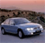 A light blue sedan with black tires and headlights is positioned on a road against a blurred background of brown terrain and a pinkish sky.