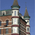 Pixelated image of a multi-story building with brown brick and white stone detailing, featuring two towers with green-topped spires against a clear blue sky.