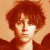 Pixelated static image of a person with curly red hair and light skin wearing dark clothing against an orange background.