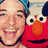 Pixelated image shows a smiling person with light skin and dark hair next to Elmo from Sesame Street, both in close-up view.
