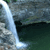 A static pixelated image of a waterfall flowing over a rocky cliff into a dark pool below, with green foliage visible at the top left corner.