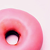 A close-up of a pink donut with a glossy surface and a dark center hole against a light background.