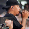 A man in a black cap and shirt sings into a microphone, his hand raised near the mic, with a smaller inset image of him singing below.