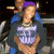 A person with purple headwear and a dark jacket over a light graphic shirt sits in front of blurred background elements.