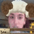 Pixelated character with light skin, brown curly hair, and a white chef's hat, wearing a yellow and brown outfit against a blurred background.