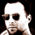 Pixelated static image of a person's face with dark sunglasses, visible facial features including nose and mouth, and brown hair on the sides against a black background.