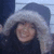 A person wearing a dark hooded coat with light fur trim smiles broadly in a snowy outdoor setting.