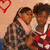 Two people embrace in a close-up pose against a light background with red heart symbols, wearing patterned and striped clothing.