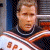 Pixelated image of a person in a high-collared uniform with orange and white stripes, standing against an interior background.