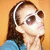 Pixelated image of a person with long brown hair wearing white sunglasses and a blue top against an orange background.