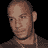 Pixelated static image of a bald man's head and shoulders with brown hair, light skin tone, wearing dark clothing against black background.
