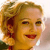 A pixelated close-up of a smiling person with light brown hair, red lips, and visible ear, set against a blurred background.