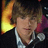 Pixelated static image of a person with brown hair wearing a white shirt and dark jacket, holding a microphone against a background featuring a yellow-orange gradient shape on the left side.