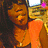 A pixelated image of a person with long dark hair wearing an orange top and necklace, smiling in front of a blurred background.
