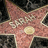 A star-shaped icon with a pinkish-red textured surface, black border, and gold trim featuring the name 'FRANK' in raised letters.