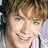A pixelated close-up of a smiling person with light brown hair holding a microphone, next to red text on a black background.