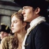 Two people in historical attire stand side-by-side, a woman with light brown hair and a man in a dark coat with white fur trim.