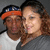 Two people pose closely together; the person on the left wears a white shirt and a blue-and-orange cap, while the person on the right wears a gray top. The background is solid gray.